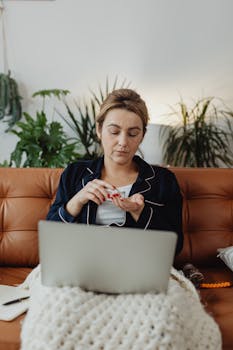 Woman in pajamas taking medicine while working on laptop from a cozy couch at home.