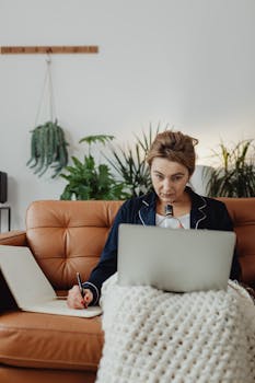 Adult woman multitasking at home using a laptop and writing in a notebook on a cozy couch.