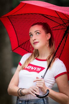 A young woman holds a stylish red umbrella while looking upward during a rainy day in New York.