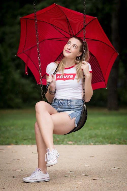 A Woman in White Shirt and Denim Shorts Sitting on a Swing with a Red Umbrella
