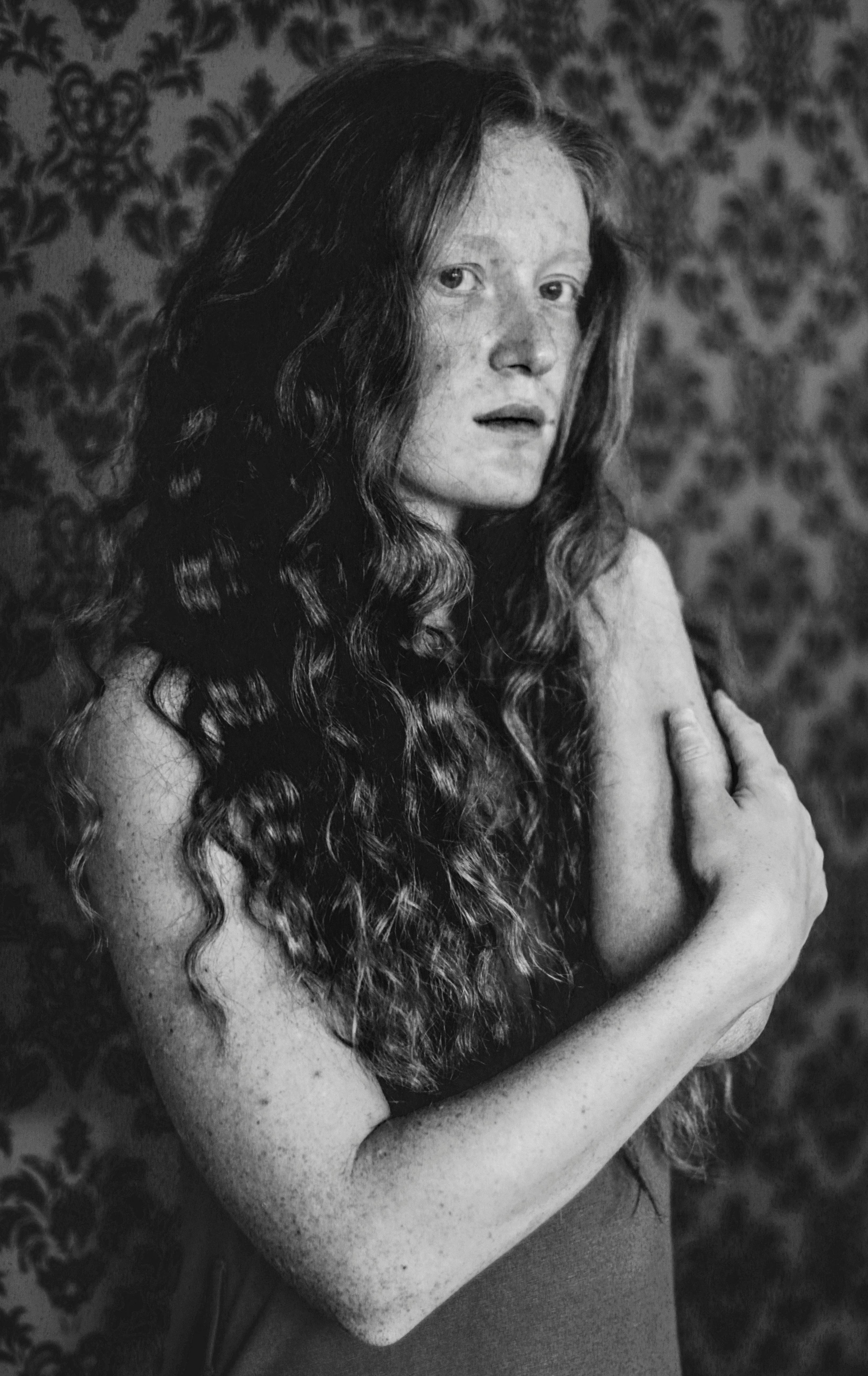 Black and white of serious female with curly hair and freckles looking at camera while standing with folded arms against wall with old fashioned design