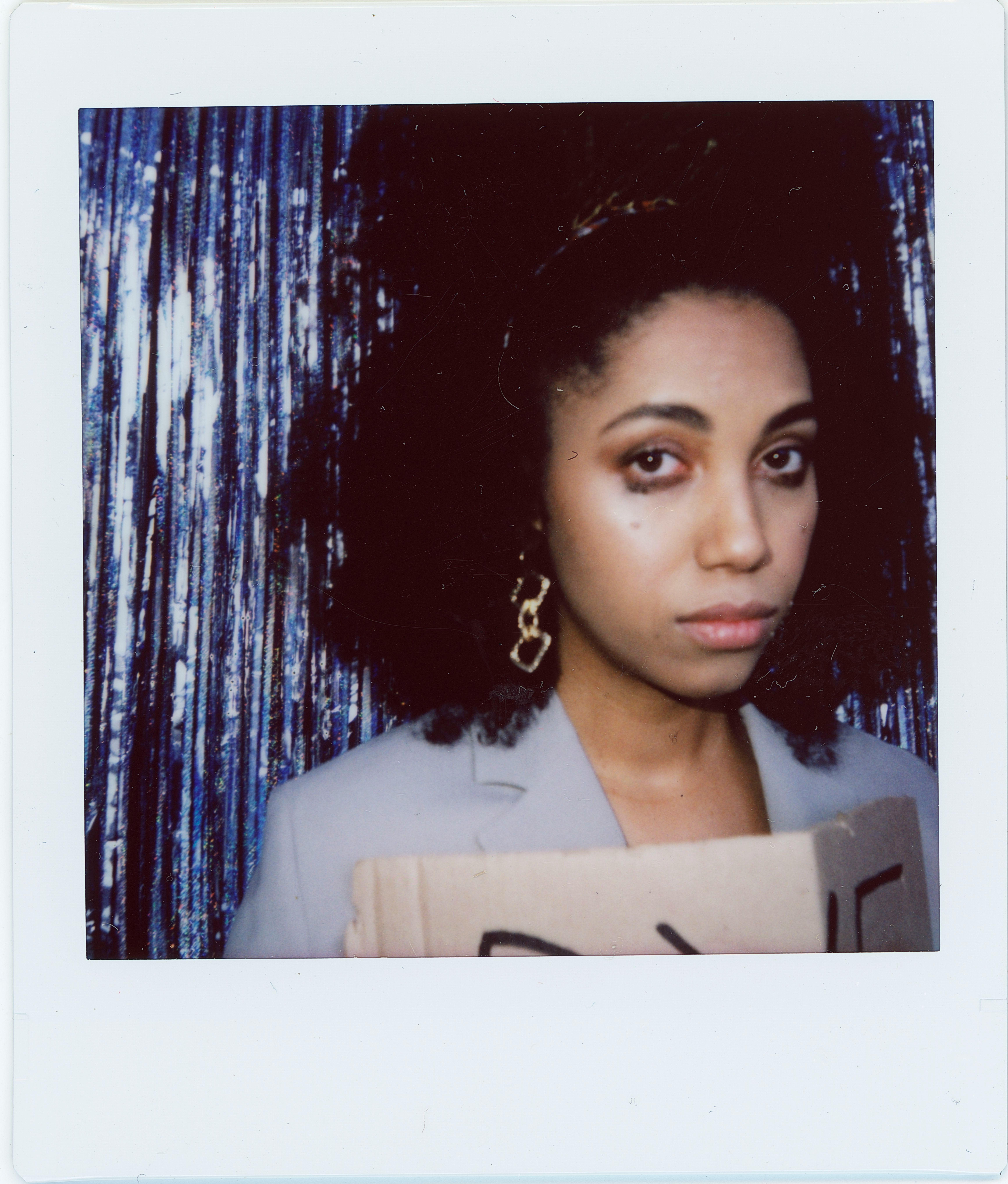 A fashionable woman with curly hair poses with a cardboard sign against a glittery backdrop. Instant photo style.