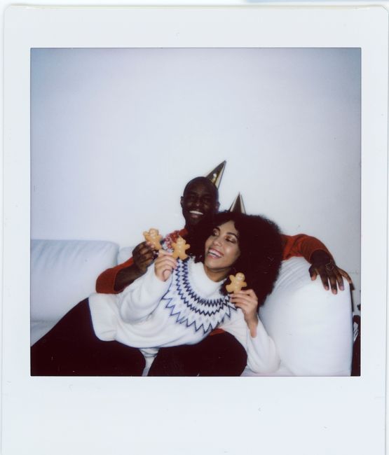 Joyful couple celebrating indoors, wearing cozy sweaters and party hats, holding gingerbread cookies.