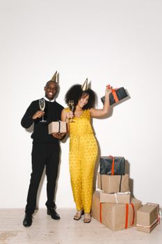 Happy couple celebrating with gifts and champagne indoors, wearing party hats.