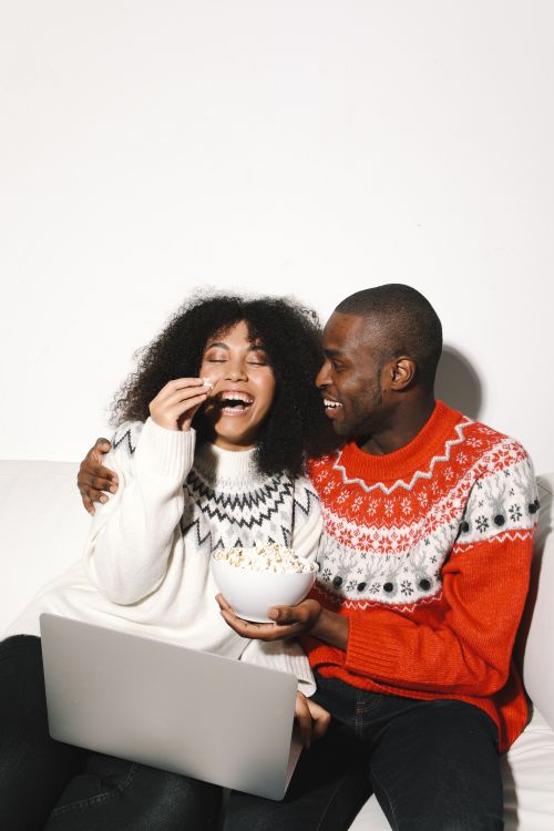 Free Smiling couple in cozy sweaters watching a movie together, enjoying popcorn on the sofa. Stock Photo