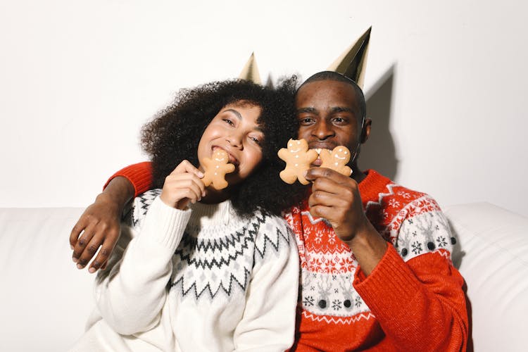 A Couple Wearing Ugly Christmas Sweaters Biting Gingerbread Cookies