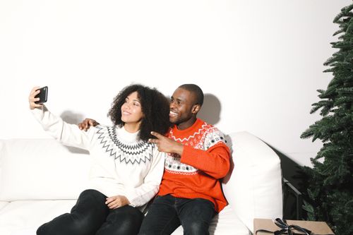 Free Smiling couple in festive sweaters taking a selfie at home with a Christmas tree. Stock Photo