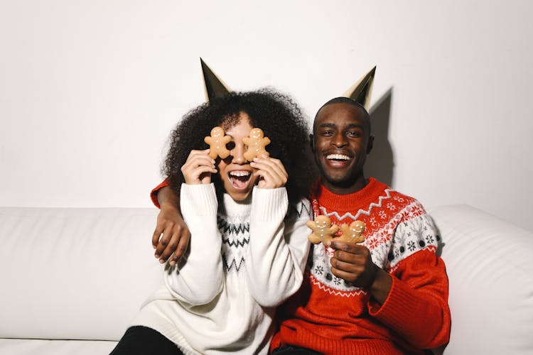 A Couple In Ugly Christmas Sweaters Holding Gingerbread Cookies