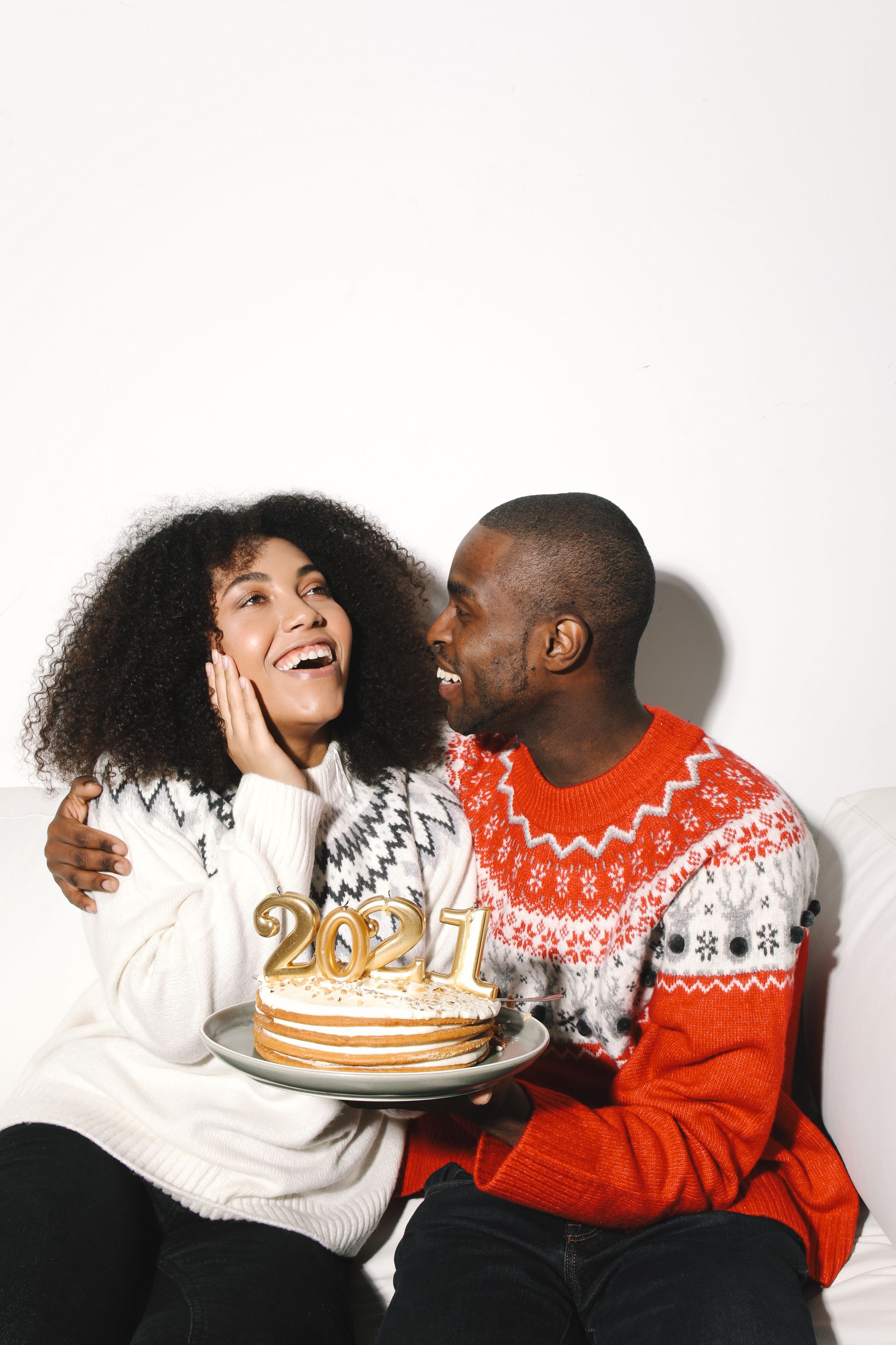 Joyful couple in winter sweaters celebrating New Year 2021 with cake indoors.