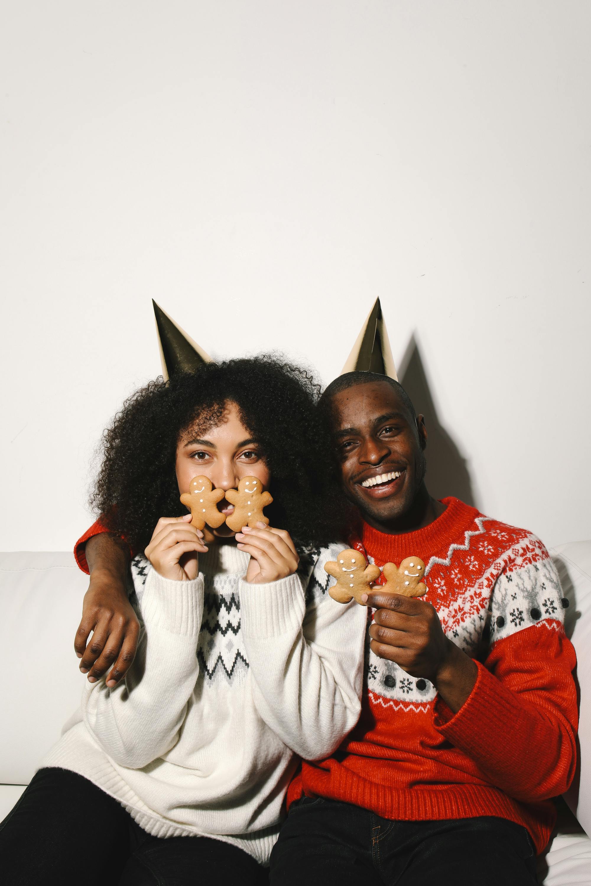 Joyful couple in festive sweaters and party hats enjoying gingerbread cookies indoors.