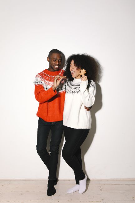 A joyful couple sharing a moment while holding a gingerbread cookie and wearing cozy sweaters indoors.