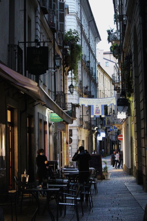 Free A narrow, atmospheric alleyway lined with cafes and storefronts in a European city at dusk. Stock Photo