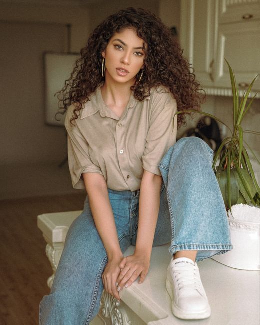 Young woman with curly hair poses stylishly in casual outfit indoors. Fashion and elegance combined.