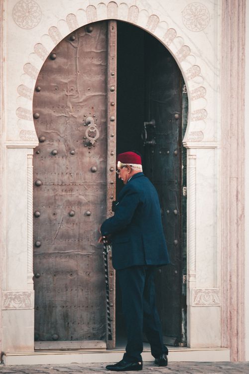 Free An elderly man in traditional attire stands by a large ornate doorway, exuding heritage. Stock Photo