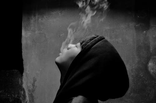 A Grayscale of a Man Wearing a Black Hoodie Blowing Smoke