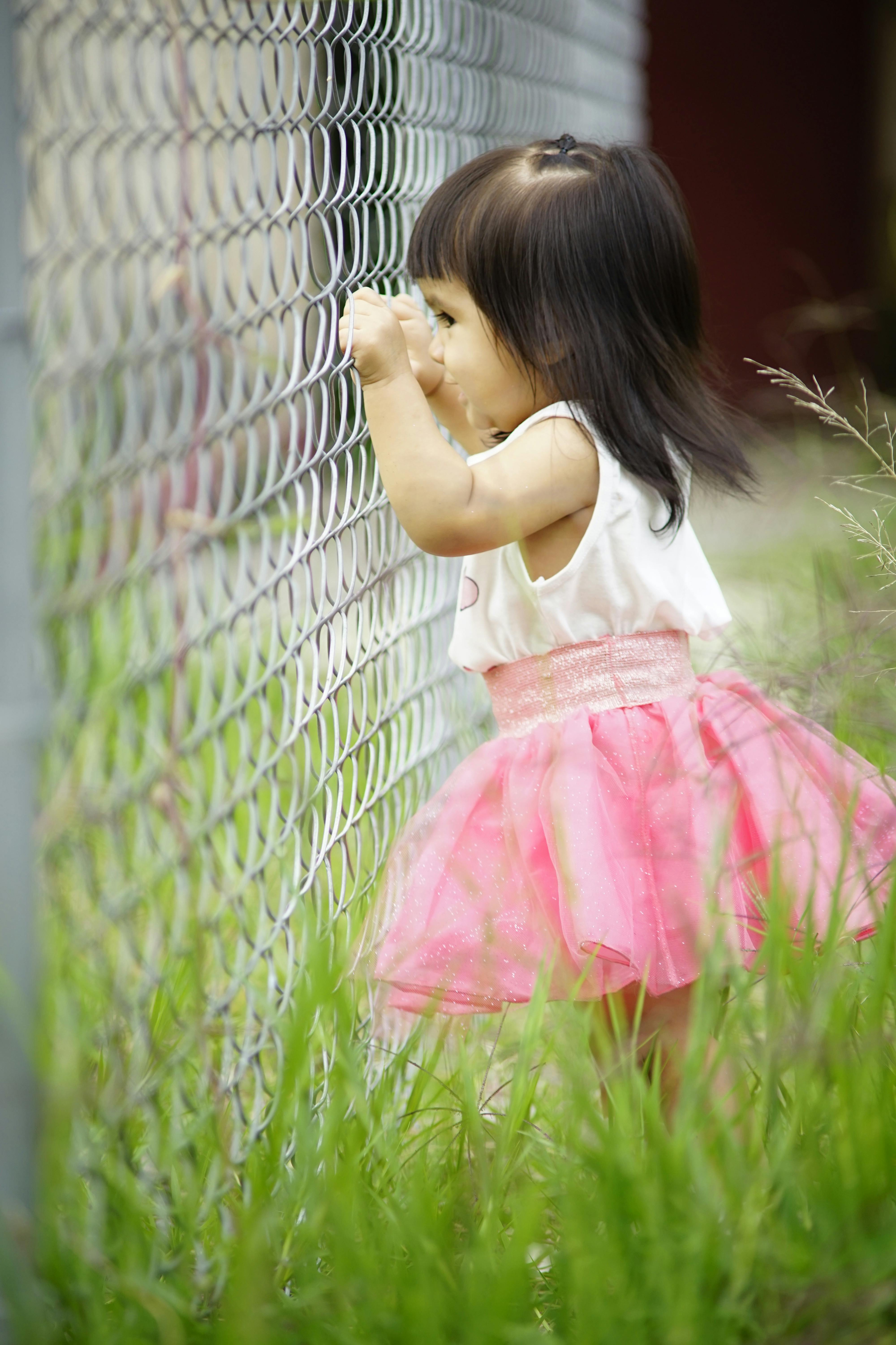 Free A young girl in a pink tutu dress looks through a wire fence in a grassy outdoor setting. Stock Photo