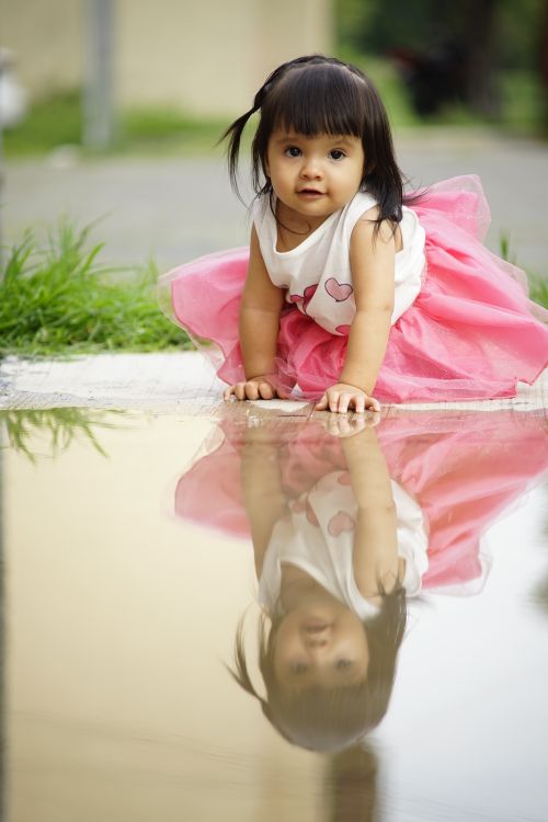 Free Sweet toddler girl in pink dress gazing at her reflection in a puddle outdoors. Stock Photo