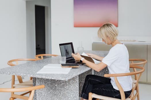 Blonde woman working remotely with laptop and notebook at a modern table indoors.