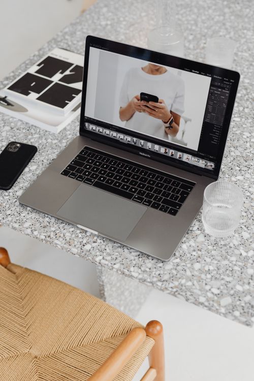 Free A laptop on a terrazzo table displaying a smartphone photo edit in a stylish workspace. Stock Photo