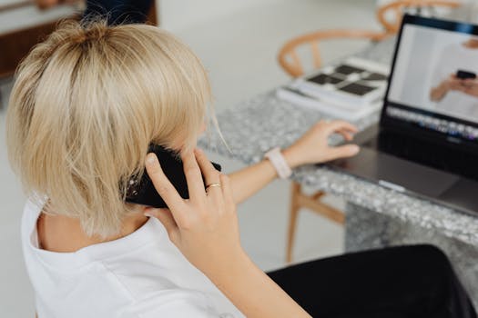Blonde woman multitasking with phone and laptop in modern workspace.