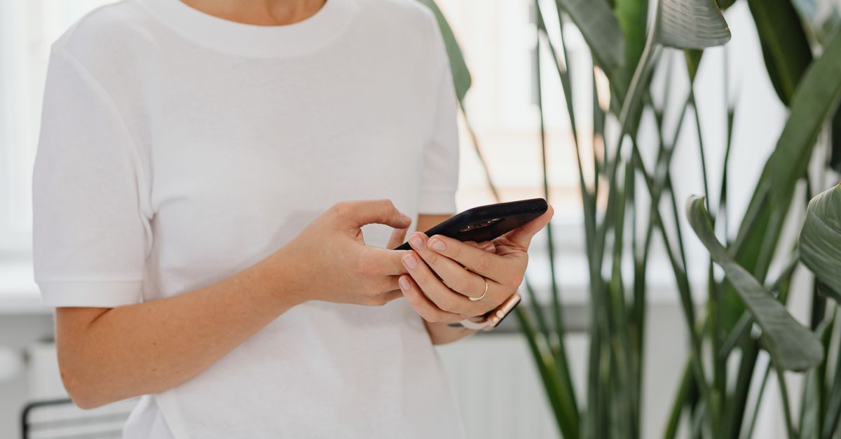 Uma mulher usa um smartphone dentro de casa rodeada por plantas verdes exuberantes.