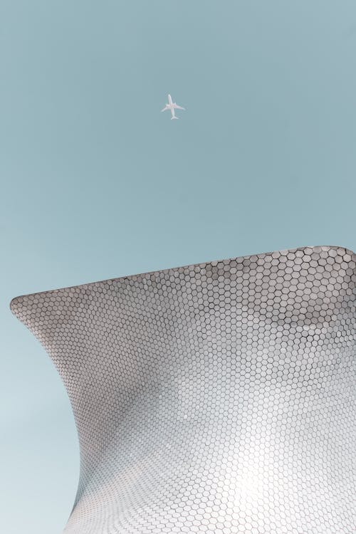 From below shot of passenger airplane flying in cloudless blue sky over modern building of unusual architecture