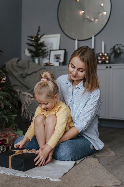 A joyful mother and daughter enjoy opening presents together beside a decorated Christmas tree indoors.