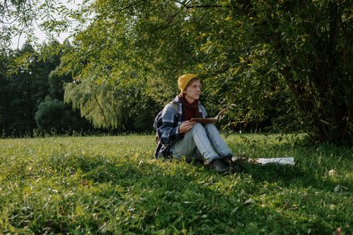 Free A man in a yellow beanie sits reading a book on a sunny day in a lush green park. Stock Photo