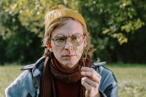 Free A young man wearing glasses and a beanie examines flora in a sunny outdoor park. Stock Photo