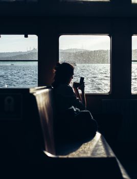 Woman on Istanbul ferry capturing skyline with smartphone through window.