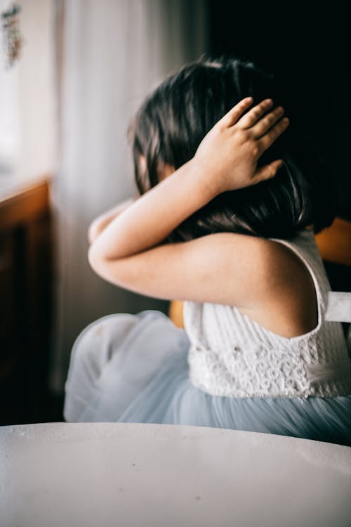 Side view of stressed unrecognizable little girl with black hair sitting at round table and touching head