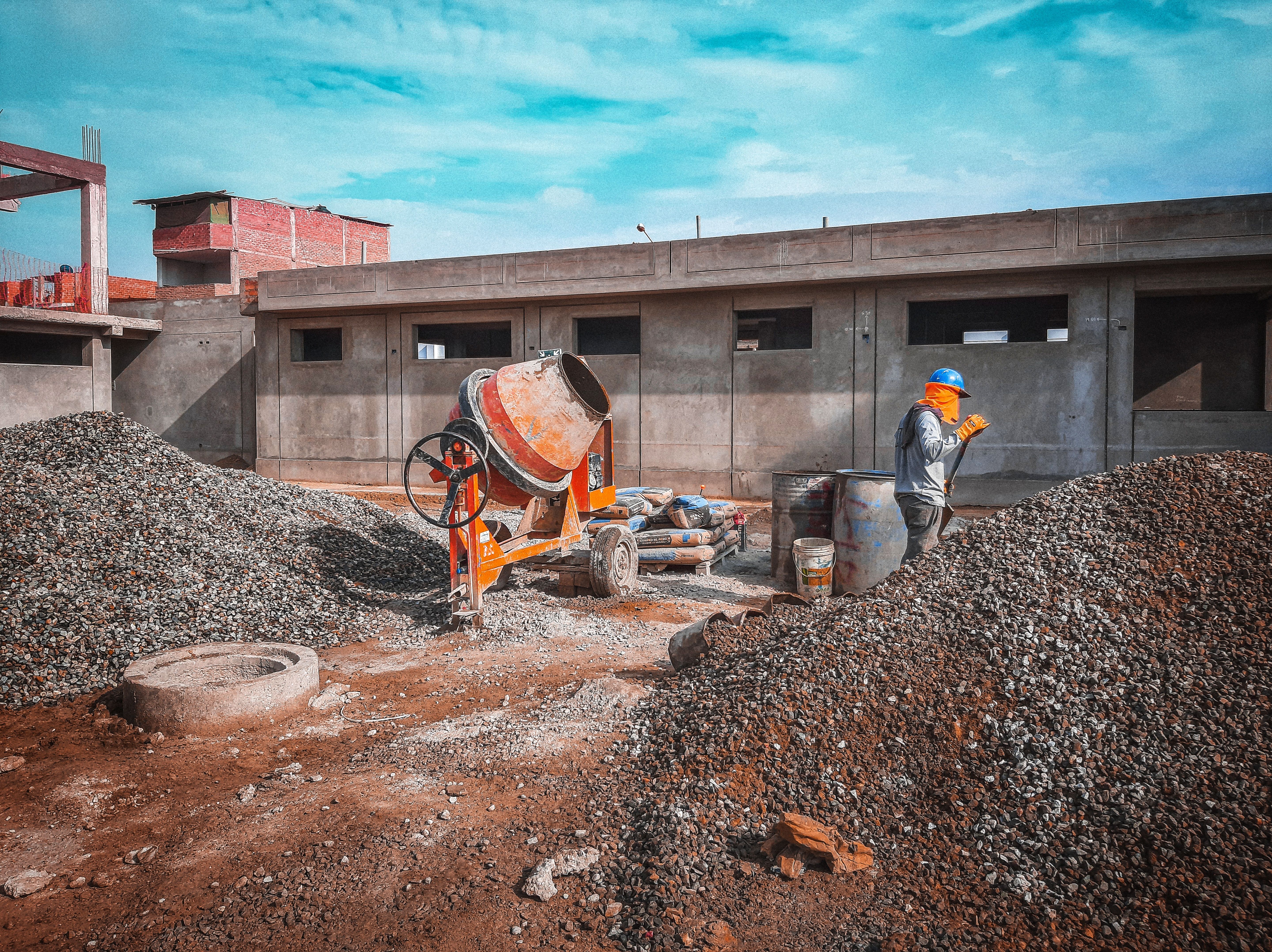 A construction site with a worke r wearing safety gear next to a cement mixer and gravel piles
