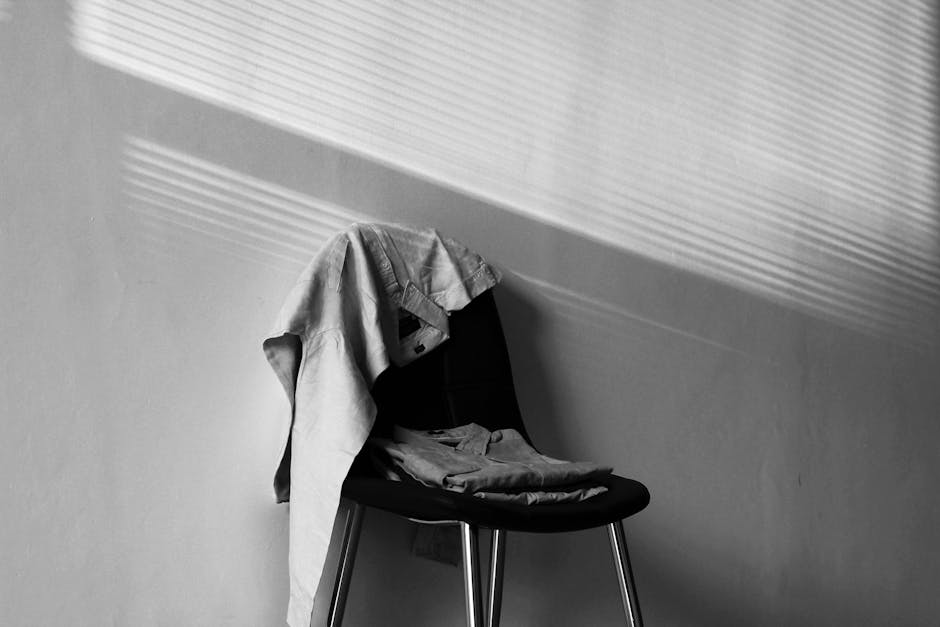 A monochrome image showcasing a chair draped with clothing under dramatic light and shadow.