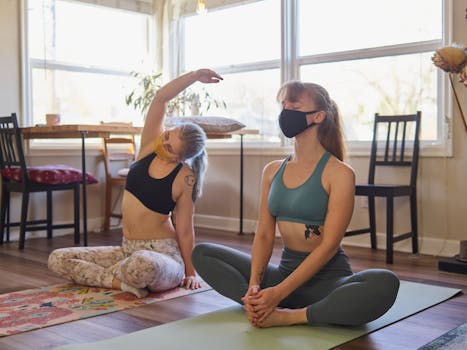 Two women practicing yoga indoors on mats, wearing face masks and workout attire for safety and relaxation.