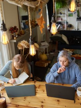 Two women studying at a wooden desk, laptops open and notebooks out, under warm lighting in a cozy home setting.