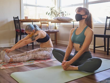 Two women practicing yoga indoors, wearing masks, focusing on wellness during the pandemic.