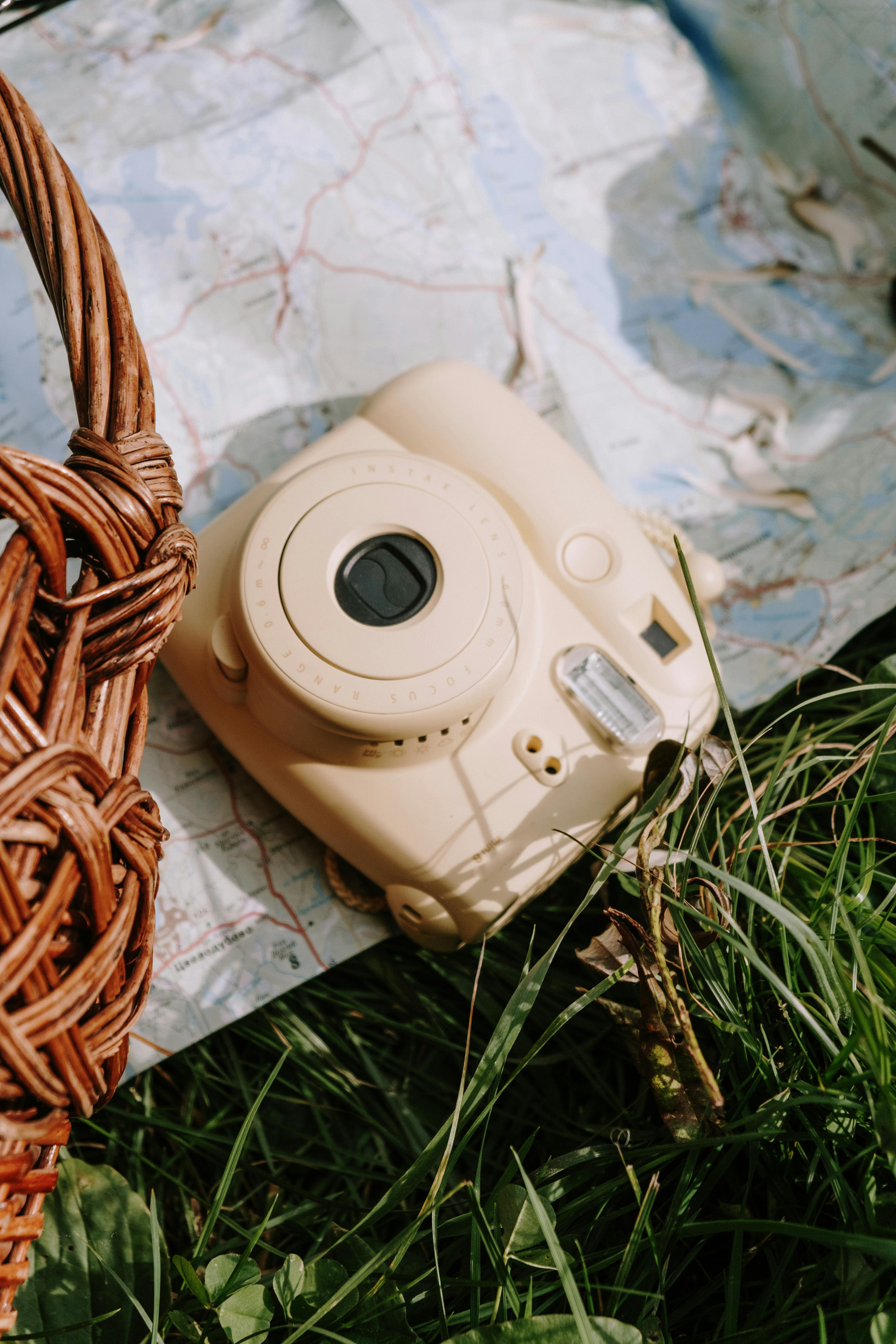 A beige Polaroid camera placed on a map outdoors, surrounded by grass and a wicker basket.