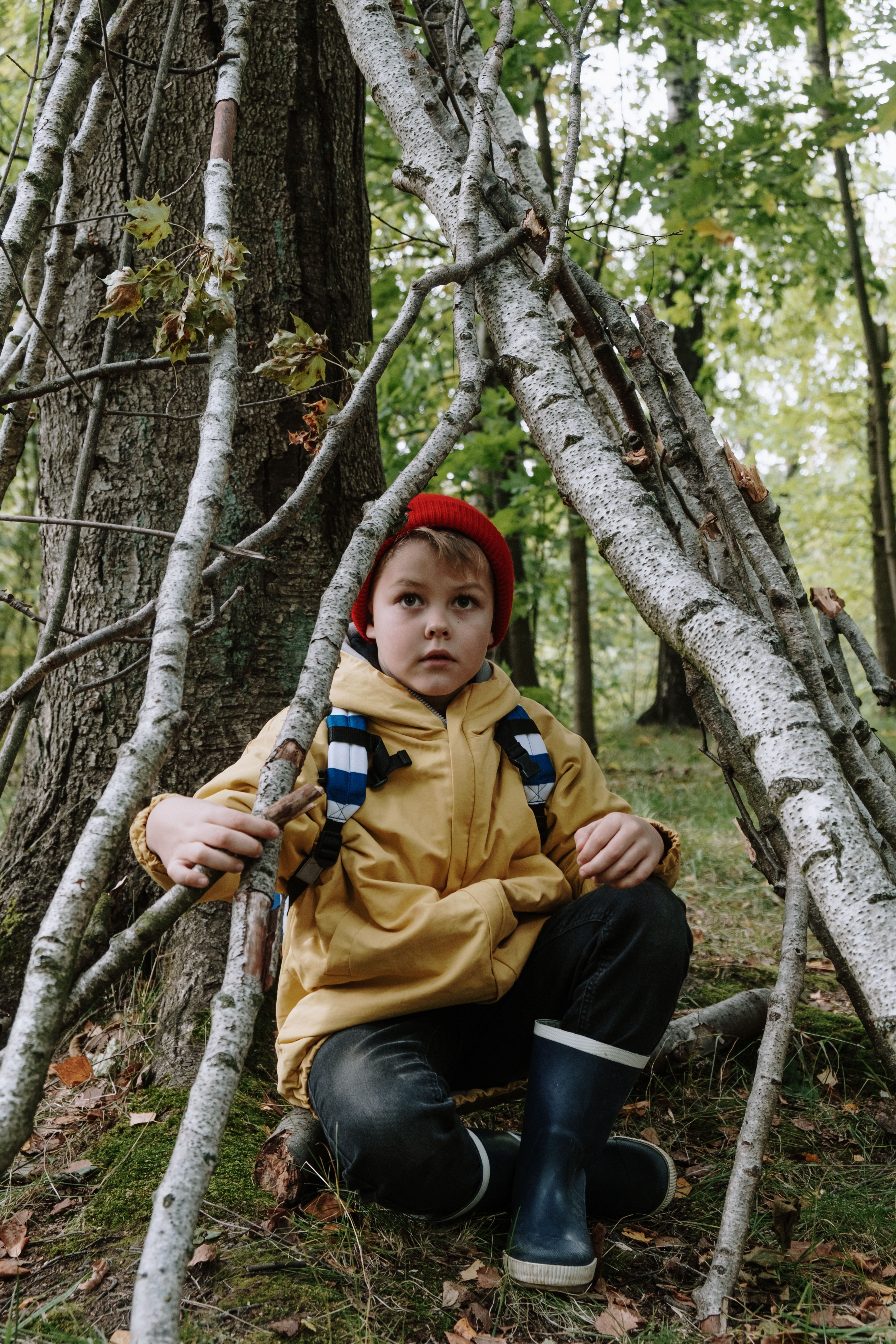 Young boy in a yellow jacket sits by a makeshift shelter in a leafy forest.