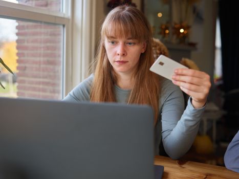 A young woman holds a credit card while using her laptop indoors.