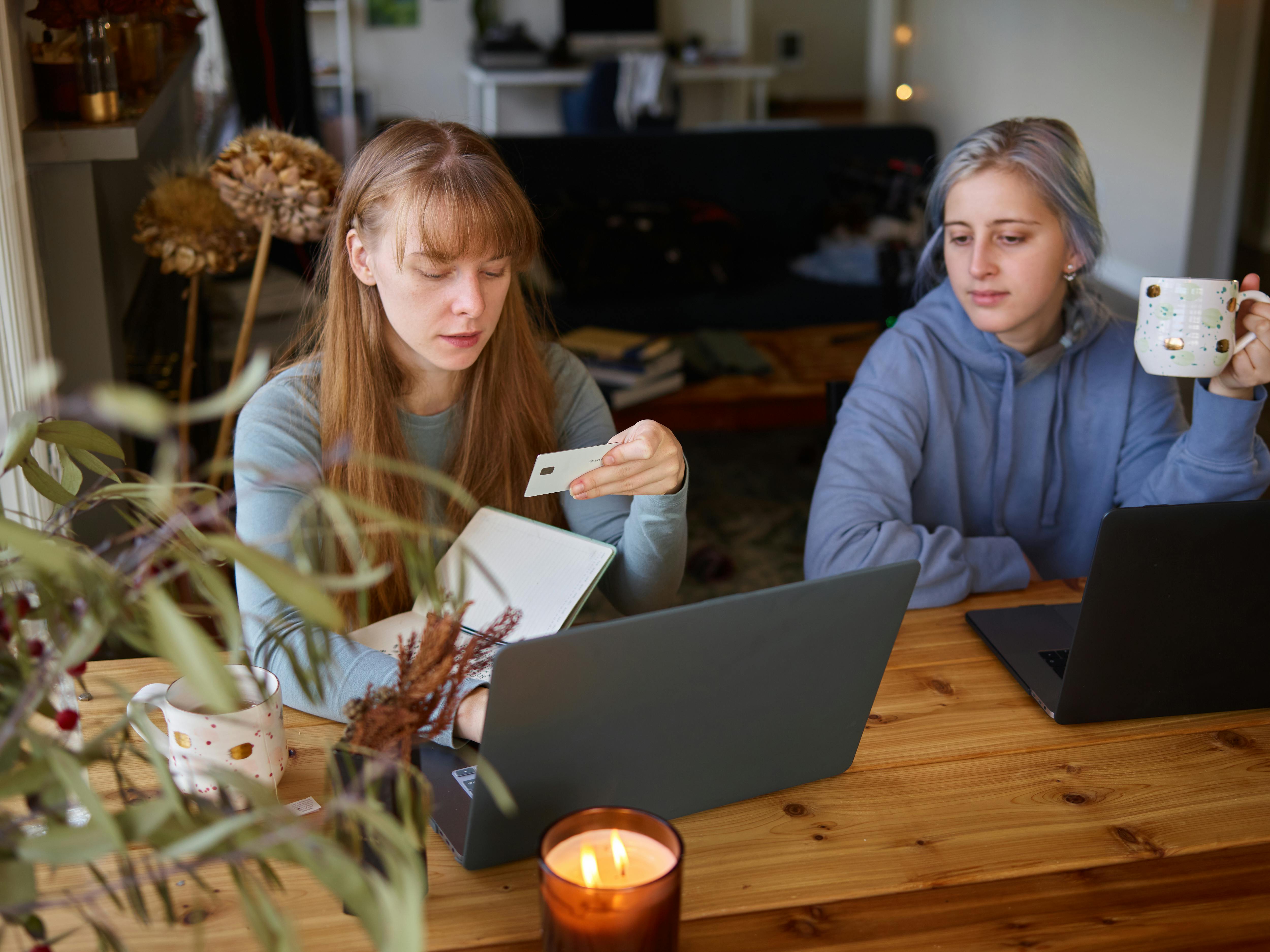 Two women using laptops for online shopping at home, holding credit cards and coffee mugs.