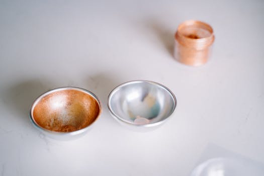 Close-up of copper and silver bowls with a glitter jar on a smooth white surface, showcasing minimalistic elegance.