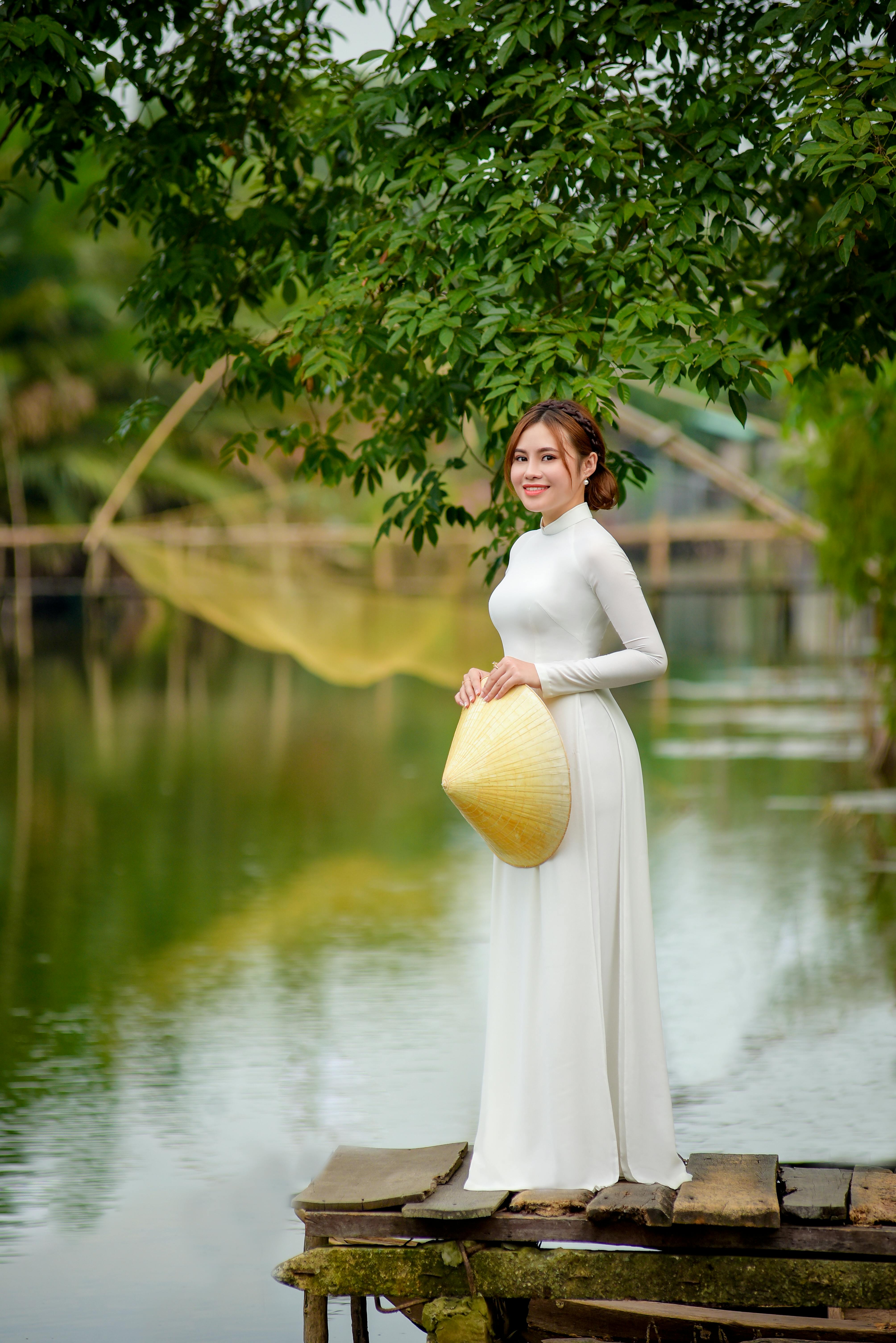 A smiling woman in a white ao dai holds a conical hat by a serene lakeside, surrounded by lush greenery.