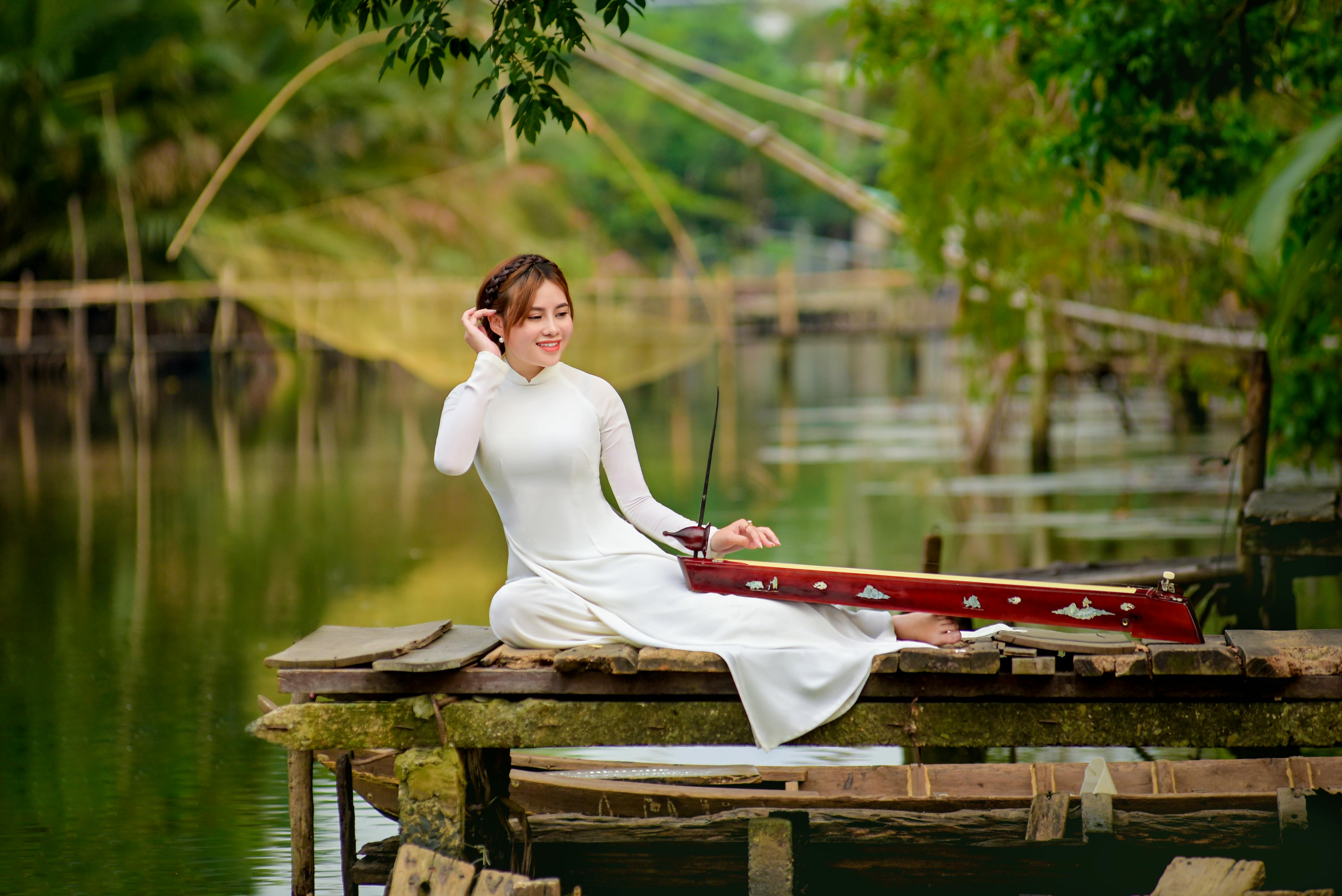 woman in white long sleeve dress sitting on brown wooden dock while holding red instrument