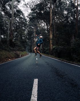 Person joyfully jumping mid-air on a winding road surrounded by tall trees in a forest area.