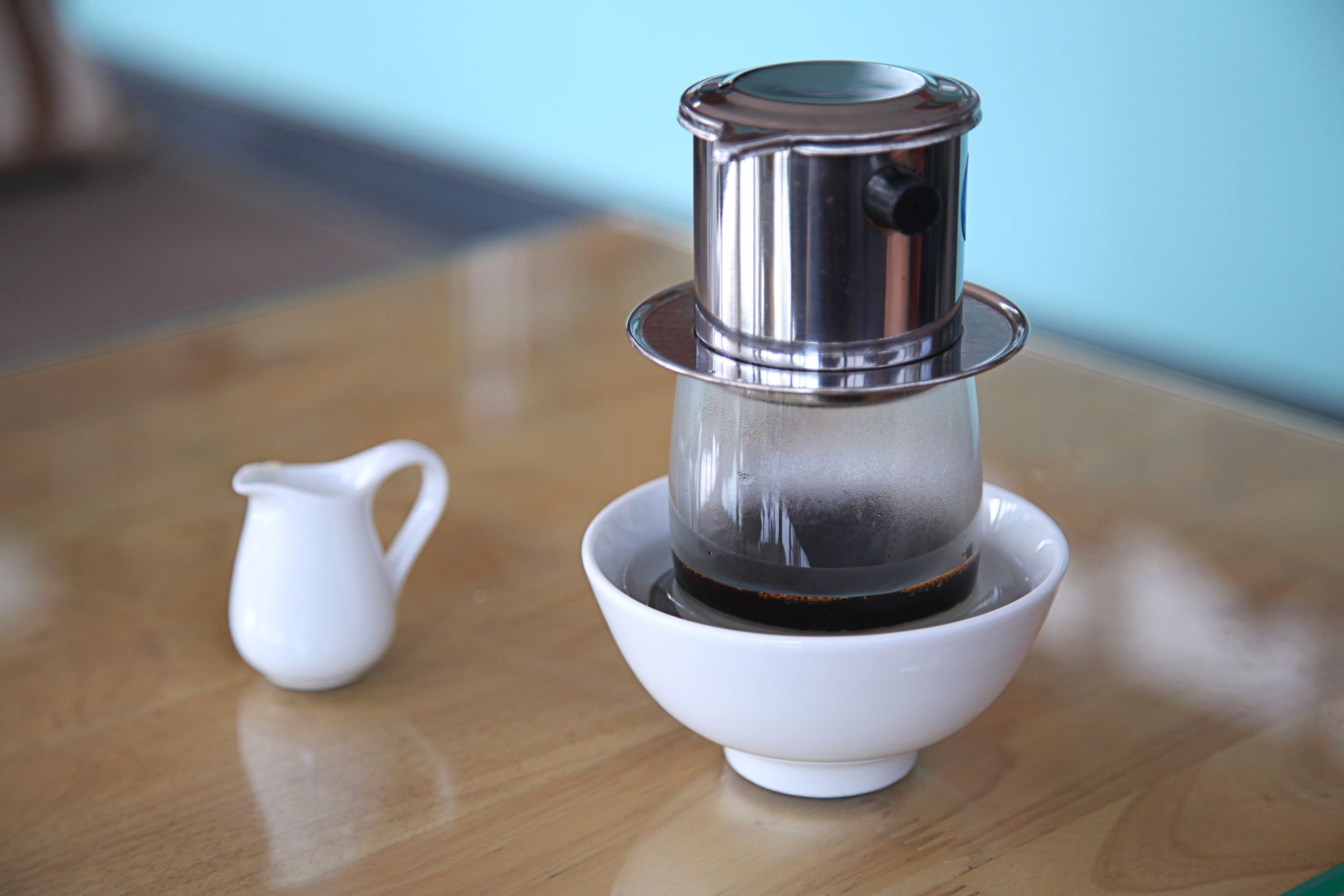 Composition of traditional Vietnamese stainless coffee filter with aromatic black coffee dripping into glass and served on wooden tables with ceramic milk pitcher