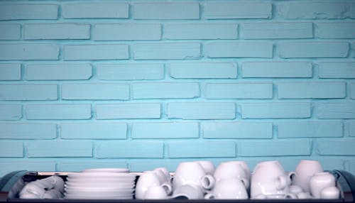 White clean ceramic plates and cups placed on table against blue brick wall after washing in kitchen