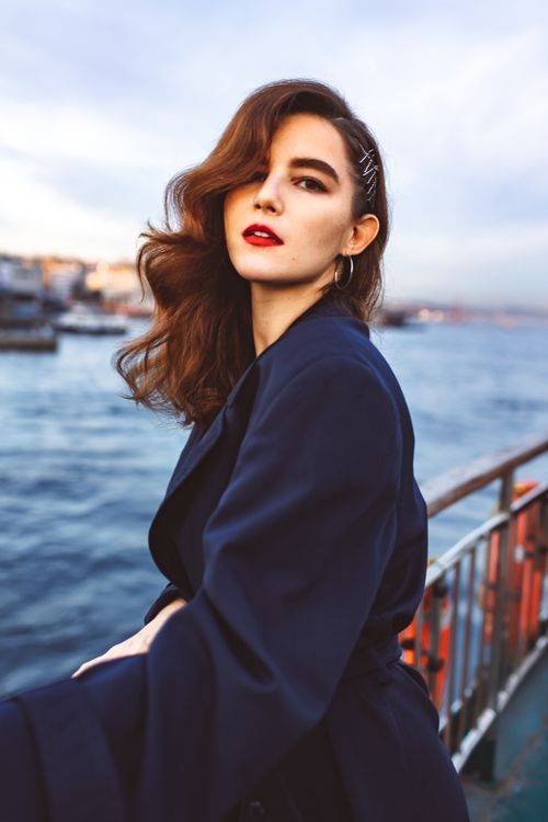 Free Stylish woman with long hair and red lipstick poses by the waterfront in a navy coat. Stock Photo