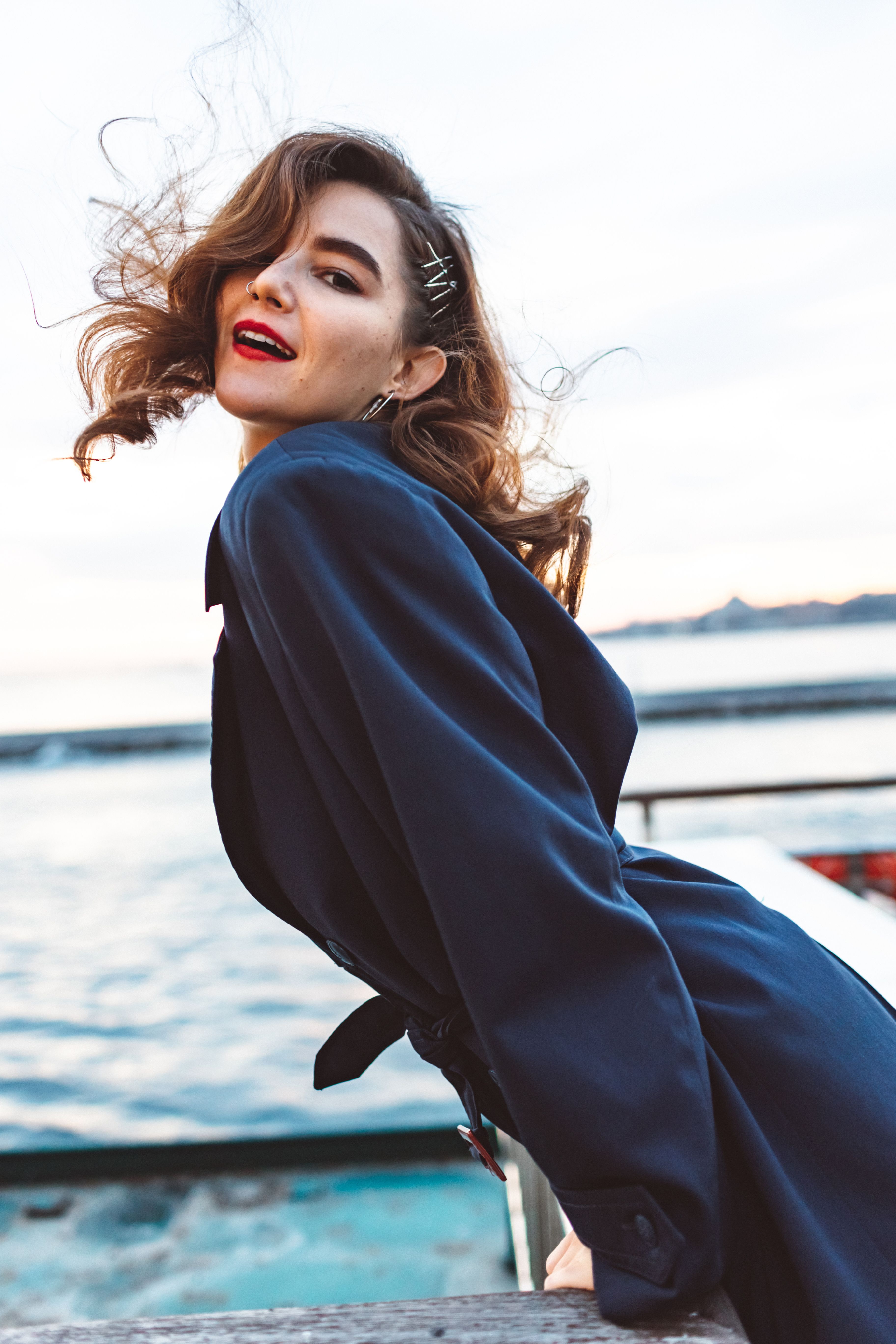 Elegant woman with curly hair and red lipstick poses by the ocean in a blue coat.