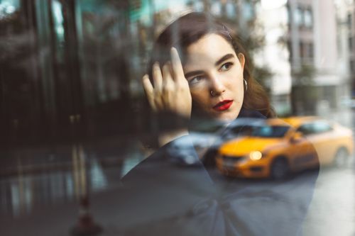 Free A young woman with red lipstick gazes through a window reflecting a bustling city street. Stock Photo