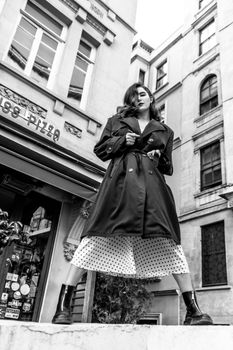 Fashionable young woman in trench coat and dress poses confidently in urban setting.